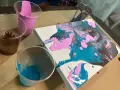 Peinture acrylique sur toile, avec des couleurs vives de rose, bleu sarcelle et marron qui coulent. Des gobelets en plastique contiennent les peintures.