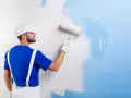 Un peintre en bleu et blanc applique une nouvelle couche de peinture bleue sur un mur blanc. C'est une peinture sur peinture en cours.