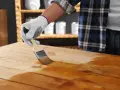Main d'une personne portant un gant blanc appliquant du vernis sur une table en bois avec un pinceau. La peinture sur bois vernis prend forme.