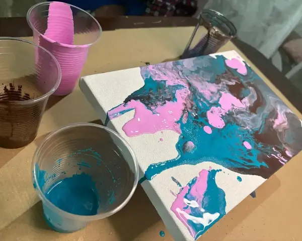Peinture acrylique sur toile, avec des couleurs vives de rose, bleu sarcelle et marron qui coulent. Des gobelets en plastique contiennent les peintures.