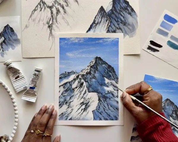 Une main peint une peinture montagne facile, avec des tubes de peinture et des nuanciers bleus à proximité.