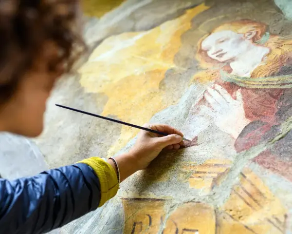 Une artiste restaure une fresque ancienne, sa main tenant un pinceau pour la préparation peinture. La scène montre une figure religieuse aux mains jointes.