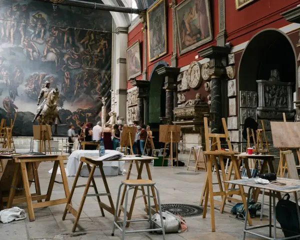 Des chevalets et des toiles dans une **école d'art appliqués**, devant une fresque monumentale et des sculptures. Des étudiants observent une œuvre.