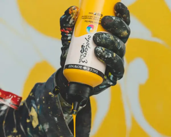 Main jaune de peinture acrylique Daler Rowney System 3, versée par une main gantée, pour un mélange de couleurs.