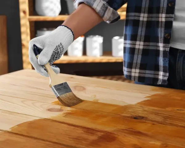 Main d'une personne portant un gant blanc appliquant du vernis sur une table en bois avec un pinceau. La peinture sur bois vernis prend forme.