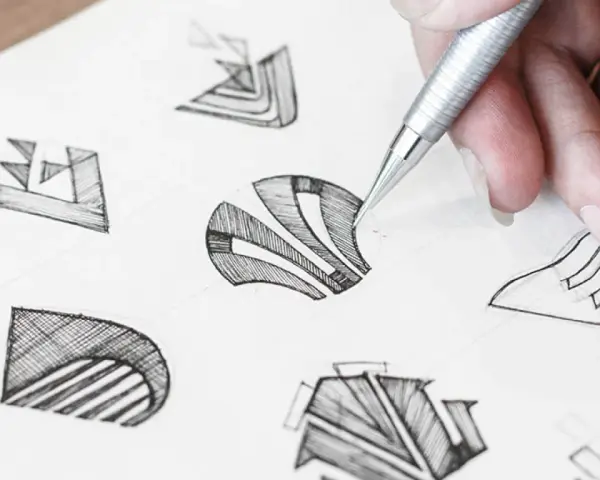 Main d'un designer esquissant des idées pour comment dessiner un logo. Plusieurs croquis abstraits sont visibles sur le papier.