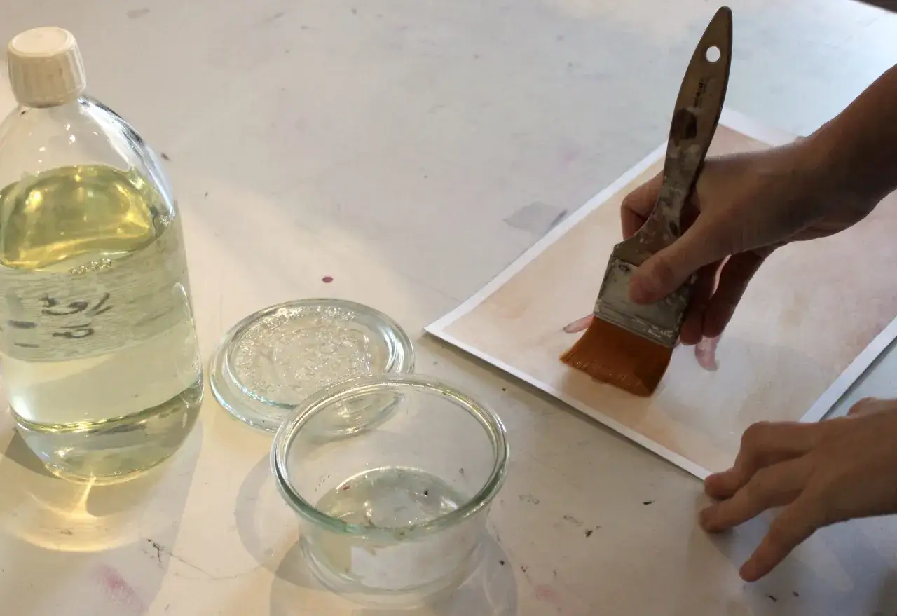 Application d'un vernis colle sur une peinture acrylique avec un pinceau. Une main maintient le papier, l'autre applique le produit.