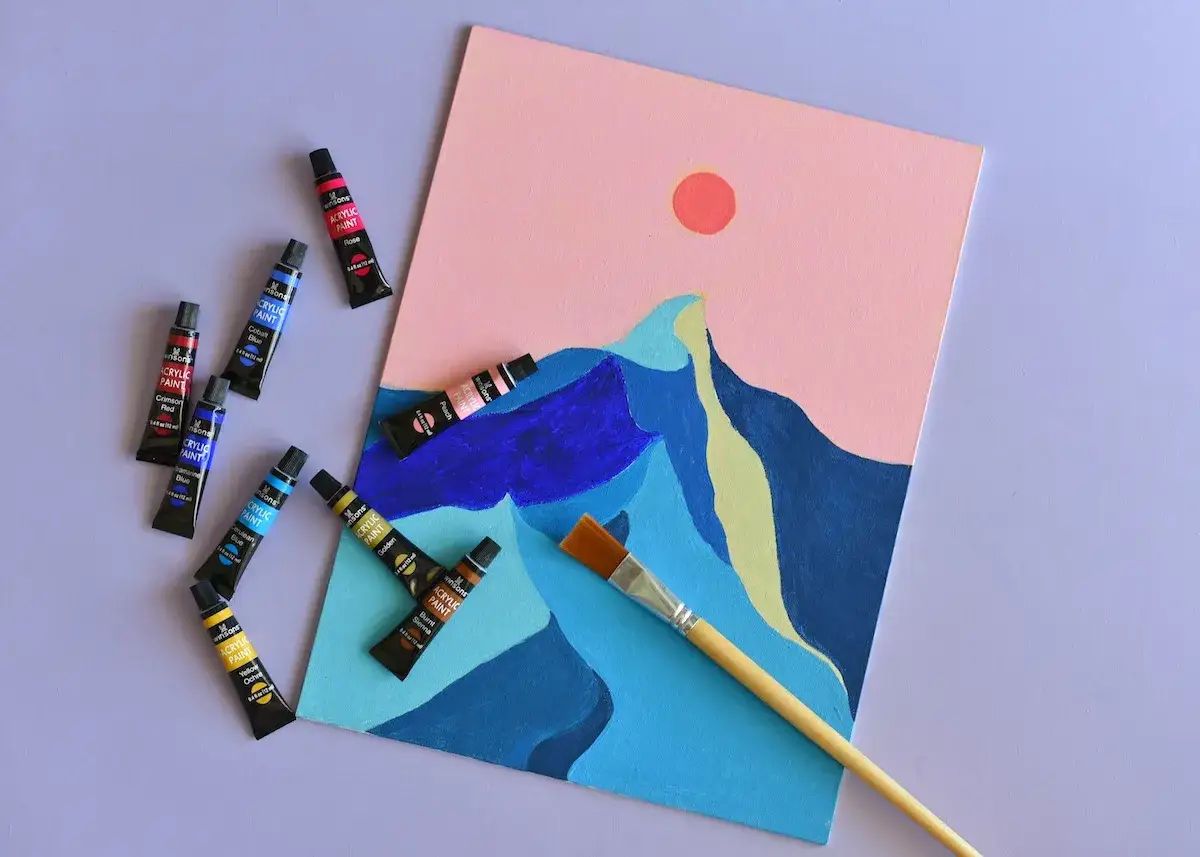 Un tableau facile et rapide à réaliser : un paysage abstrait de montagnes bleues sous un ciel rose avec un soleil orangé. Des tubes de peinture acrylique et un pinceau sont disposés autour.