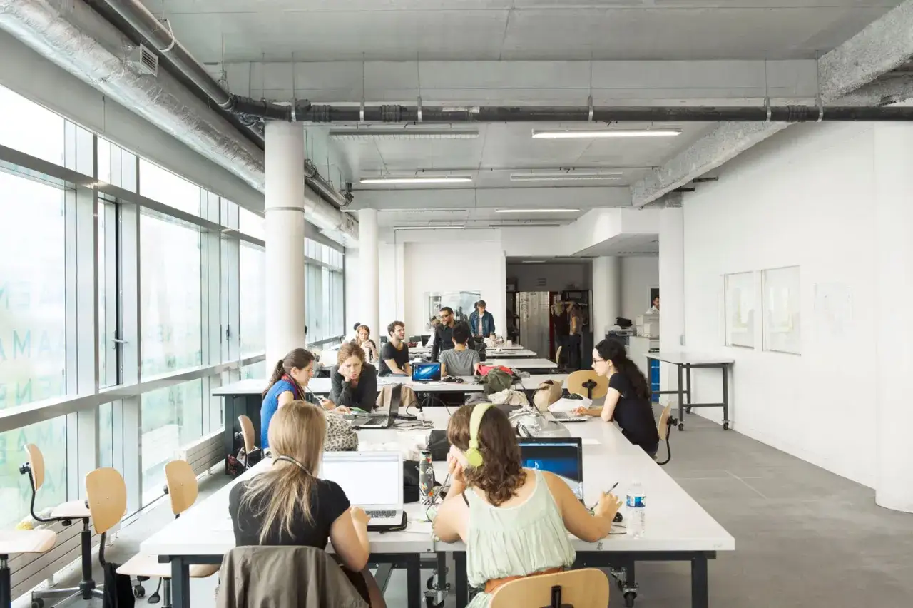 Des étudiants travaillent dans un espace ouvert, peut-être une école d'art, avec des bureaux et des ordinateurs. Le ensaama classement est visible sur les vitres.
